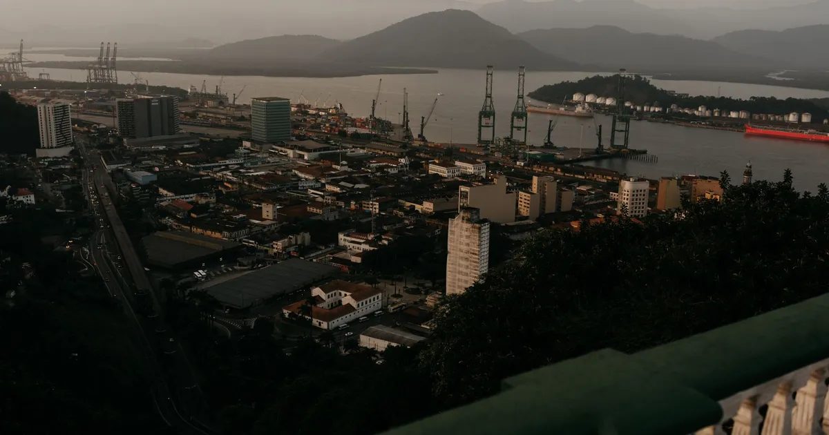 Porto com guindastes de carga em cidade costeira, simbolizando o agronegócio e comércio internacional brasileiro em busca ...