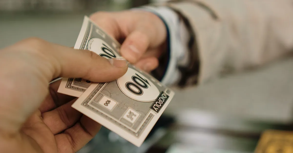 Mãos trocam dinheiro Monopoly, simbolizando acordo Braskem, BTG Pactual e proventos BradSaúde.