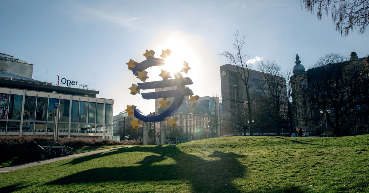 Escultura do Euro em Frankfurt, Alemanha, com paisagem urbana; reflete juros estáveis Europa e atenção à atividade econômica.