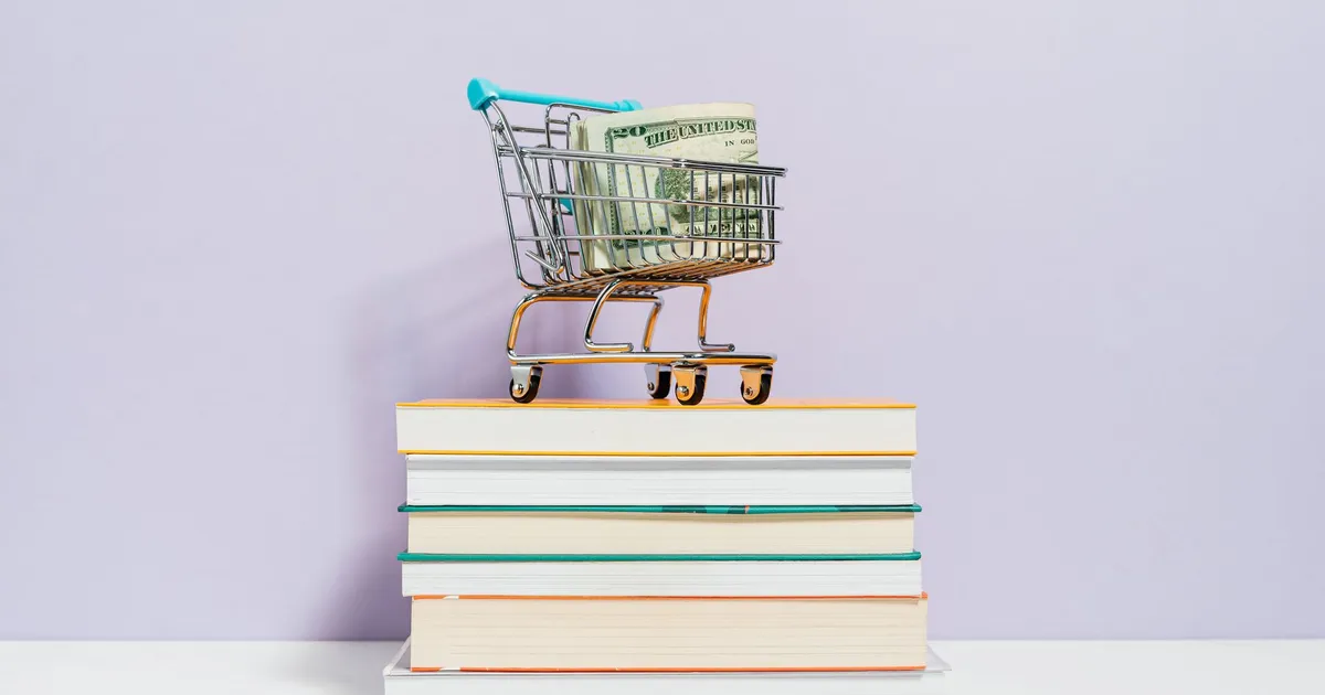 Carrinho de compras com dólares sobre livros, representando consumo e finanças em meio a juros e incertezas.