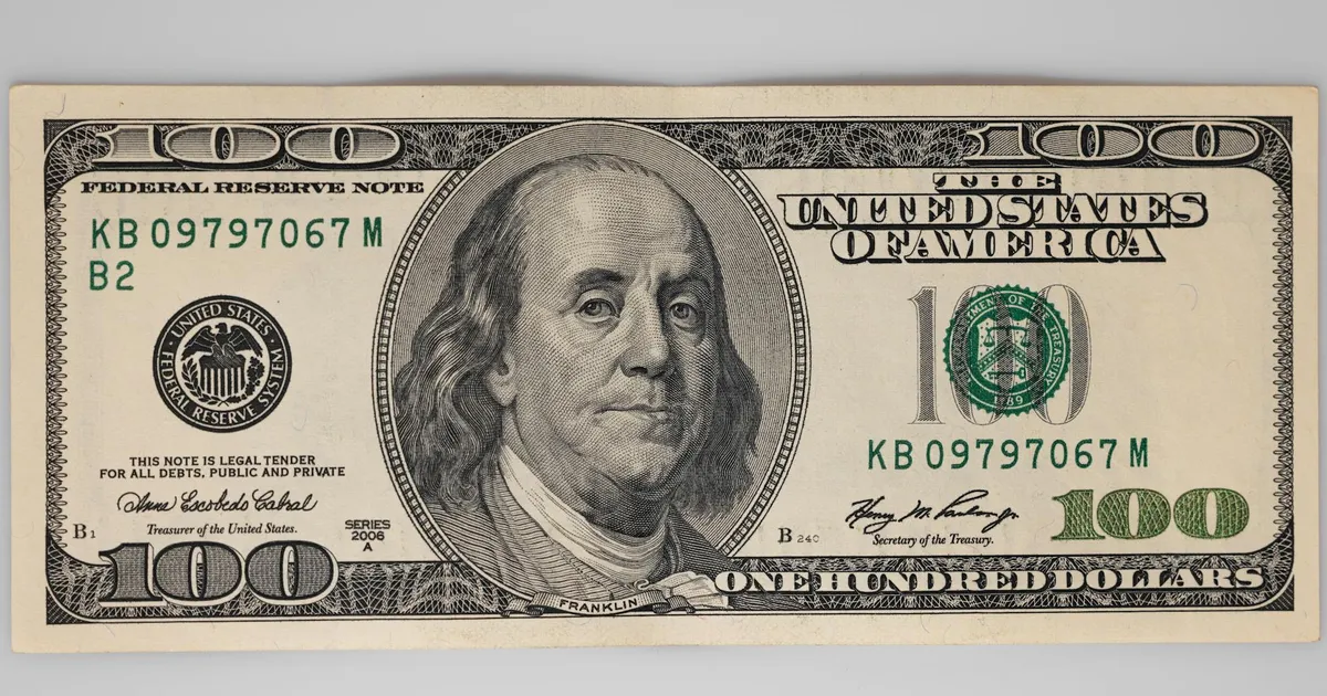 Nota de 100 dólares com Benjamin Franklin. Juros nos EUA, dólar e inflação em debate.