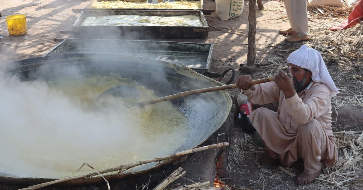 Homem prepara suco de cana em panela gigante, representando produção de açúcar na Índia e seu impacto no mercado.
