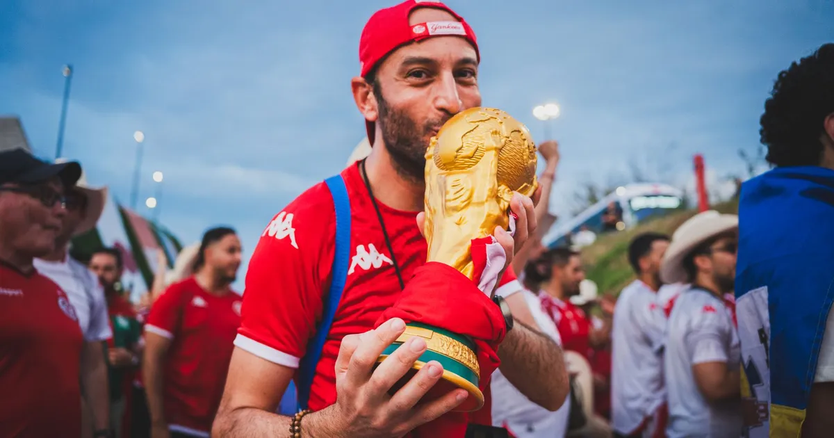 Torcedor celebra troféu da Copa 2026; artigo discute prêmios em dólar e câmbio para seleções.