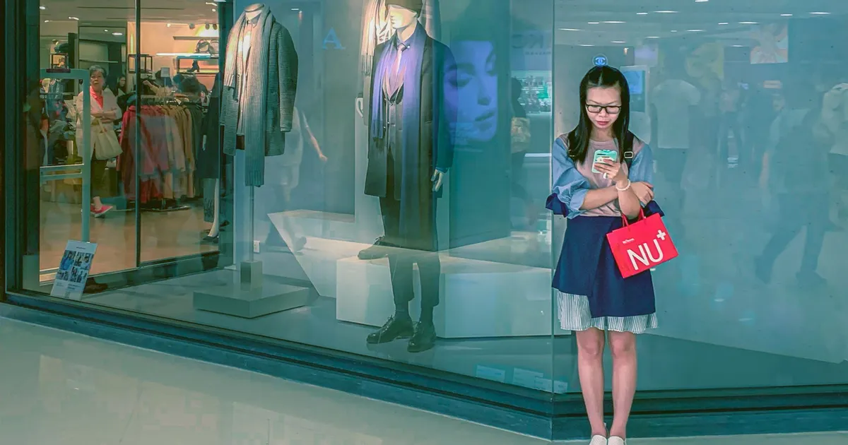 Shein IPO: Jovem asiática com sacolas de compras no shopping foca em celular, refletindo o impacto do varejo de moda.